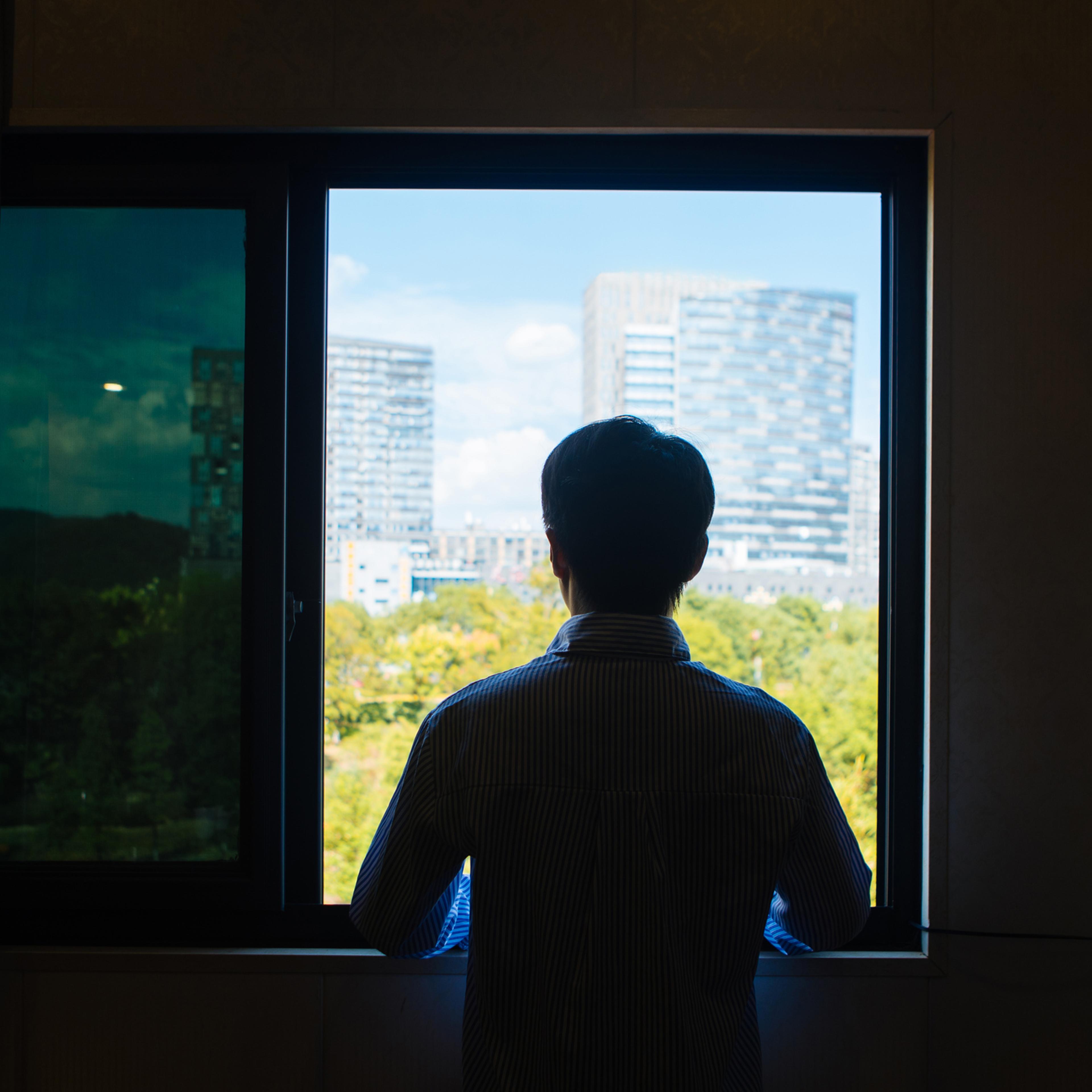 Young man in China standing in front of a window looking into the distance, with a green park and modern skyscrapers outside.