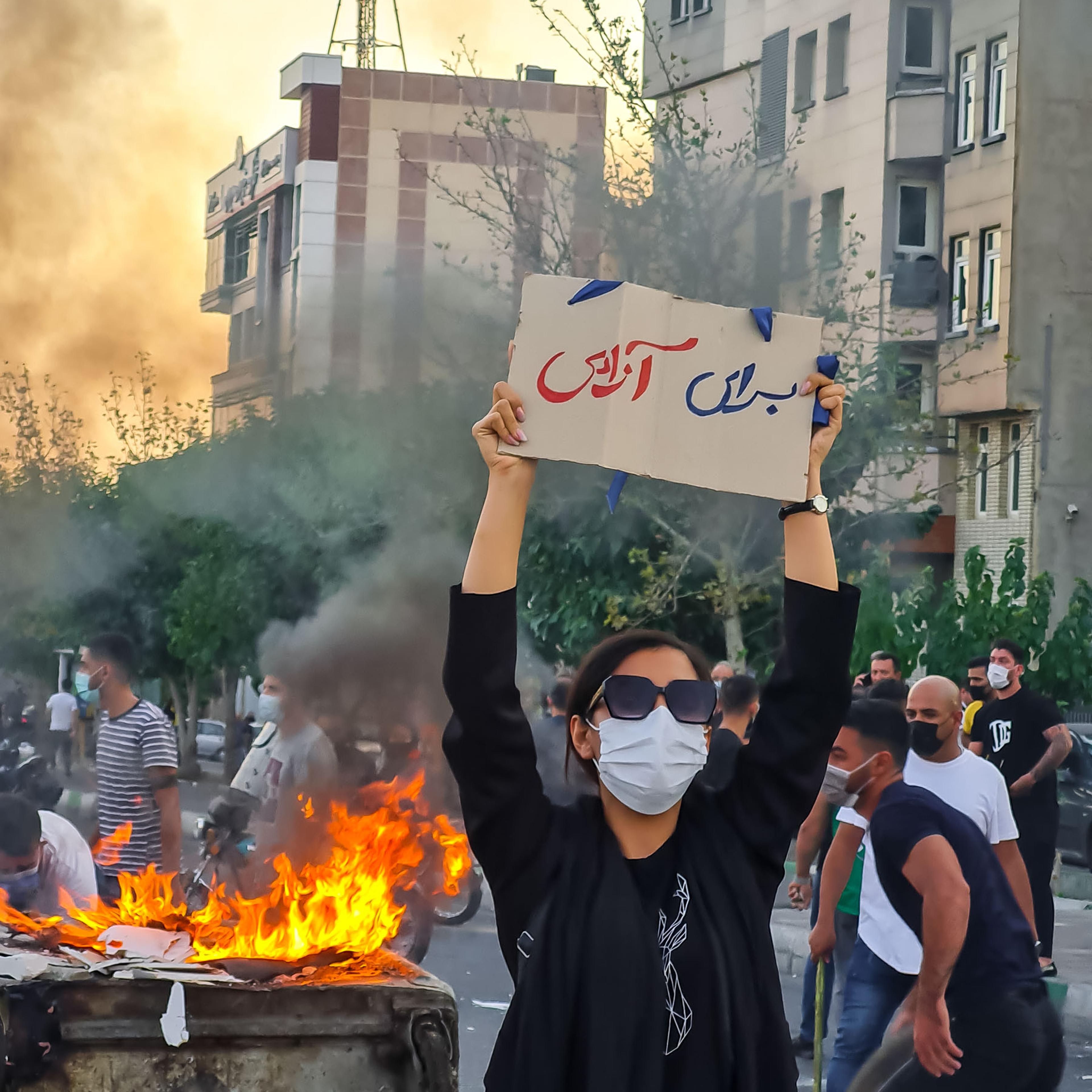 A woman without hijab, holding a sign written on it "For Freedom" standing next to a burning dumpster, with trashes on fire, protesting against the Islamic Republic's mandatory hijab law and the death of Mahsa Amini, in the western part of Tehran, Iran.