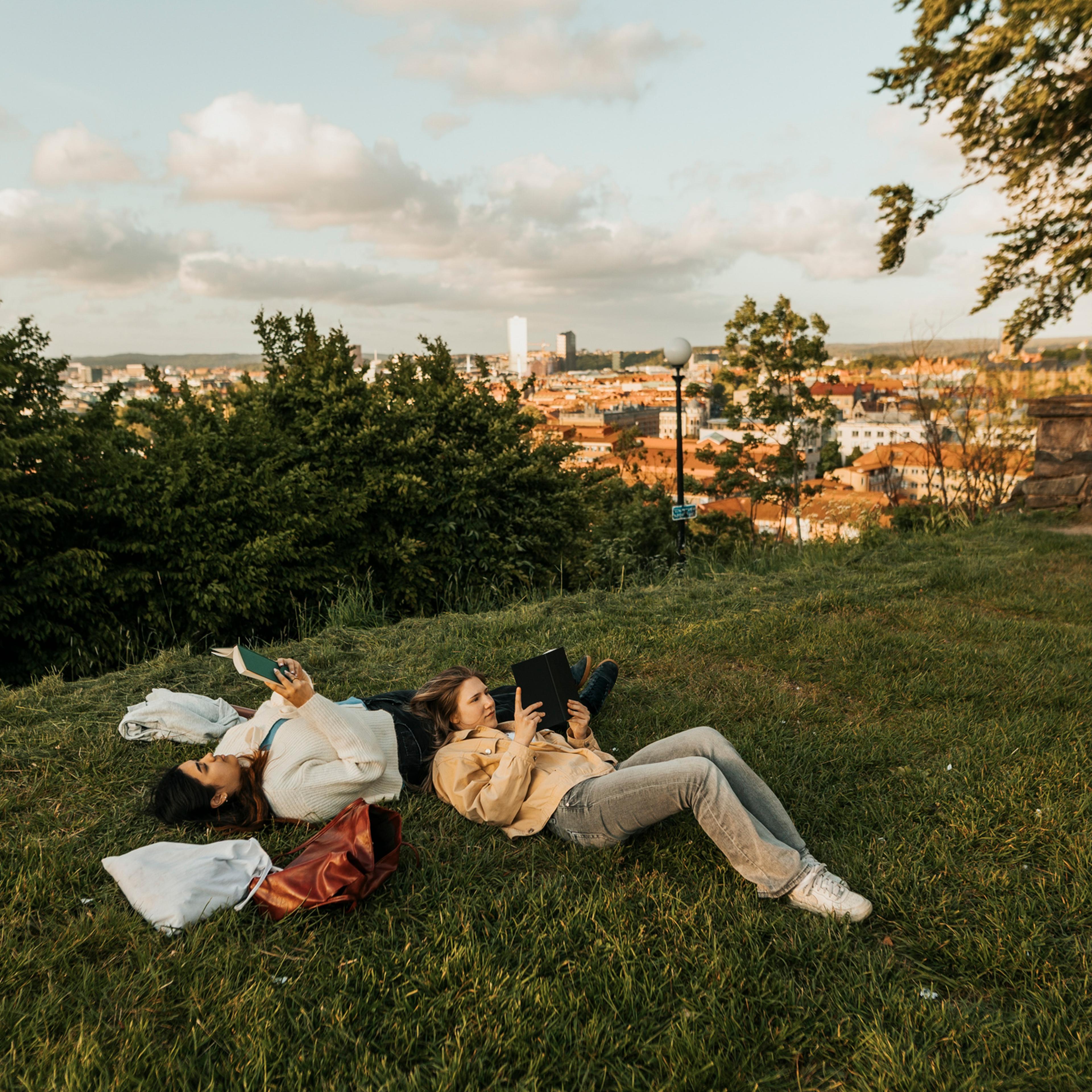 Female friends reading book while lying down on grass at a hill.