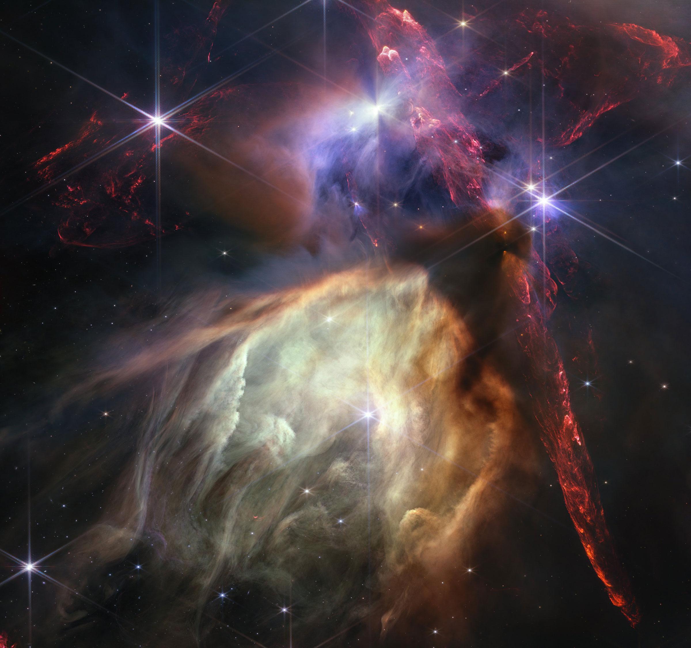 Red dual opposing jets coming from young stars fill the darker top half of the image. At bottom center is a glowing pale yellow, cave-like structure, its top tilted toward two o’clock, with a bright star at its center. The dust of the cave structure beco