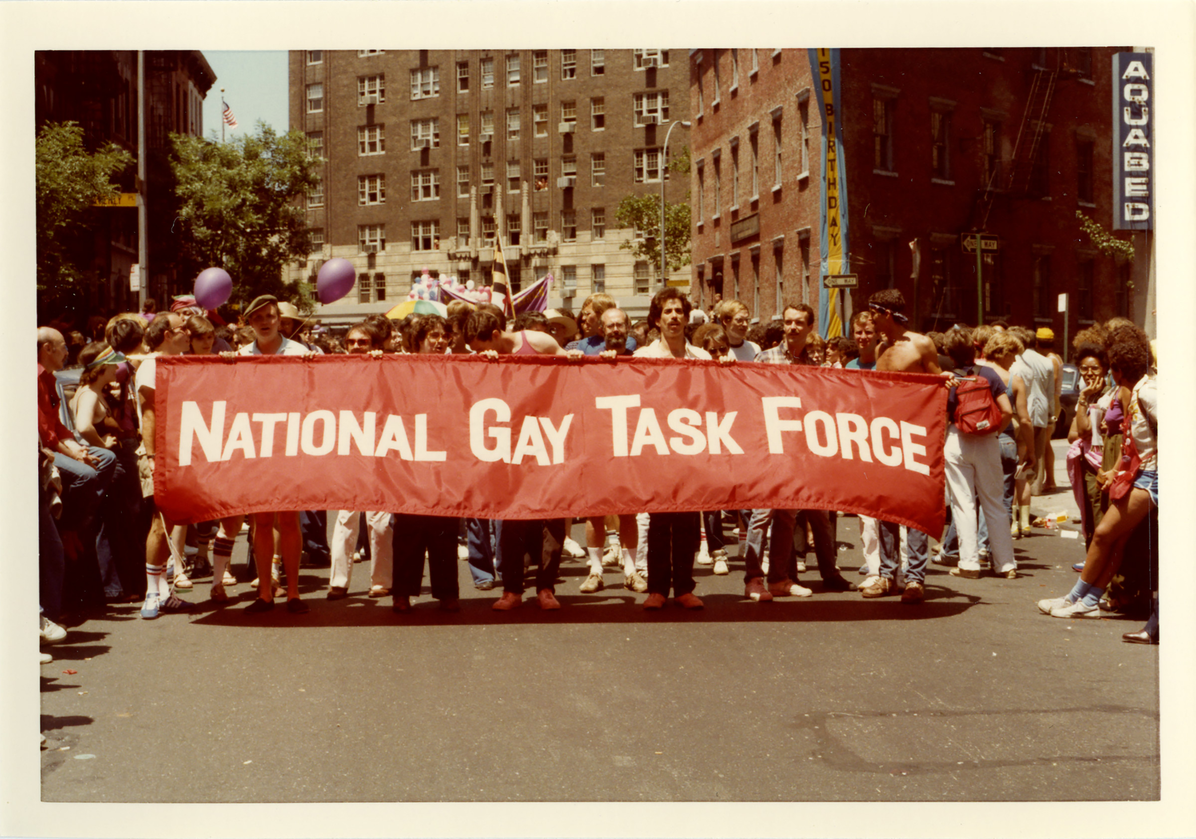 A vintage photograph from an early LGBTQ pride march, capturing a group of activists walking down a city street while holding a large red banner that reads "National Gay Task Force" in white block letters. Spectators and historic brick apartment buildings line the background.