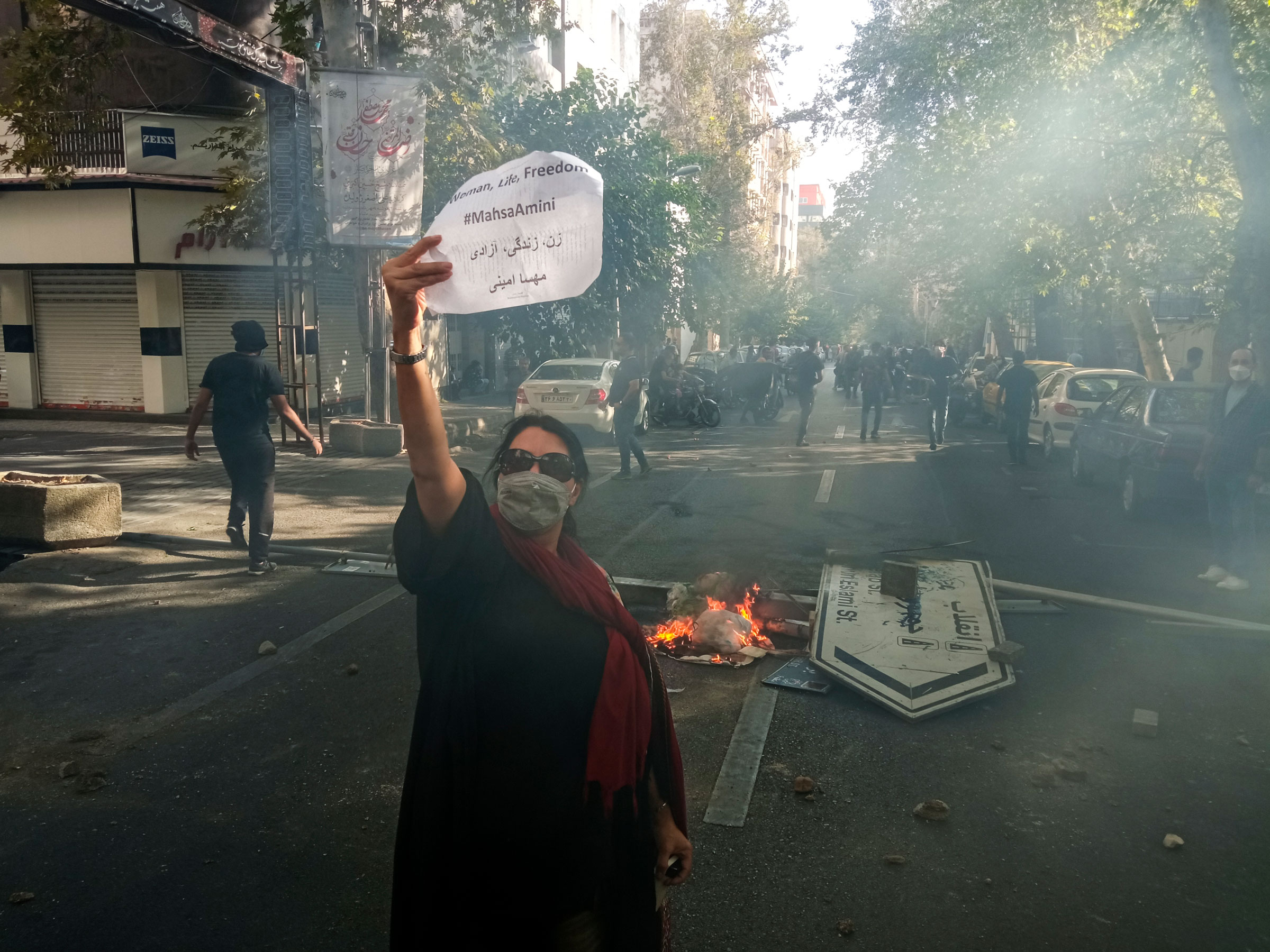 A woman holding the "Woman, Life, Freedom" sign, with the fallen street sign "Jomhouri Eslami" (Islamic Republic) in the background and tear gas in the air. October 1st 2022. Tehran, Iran.