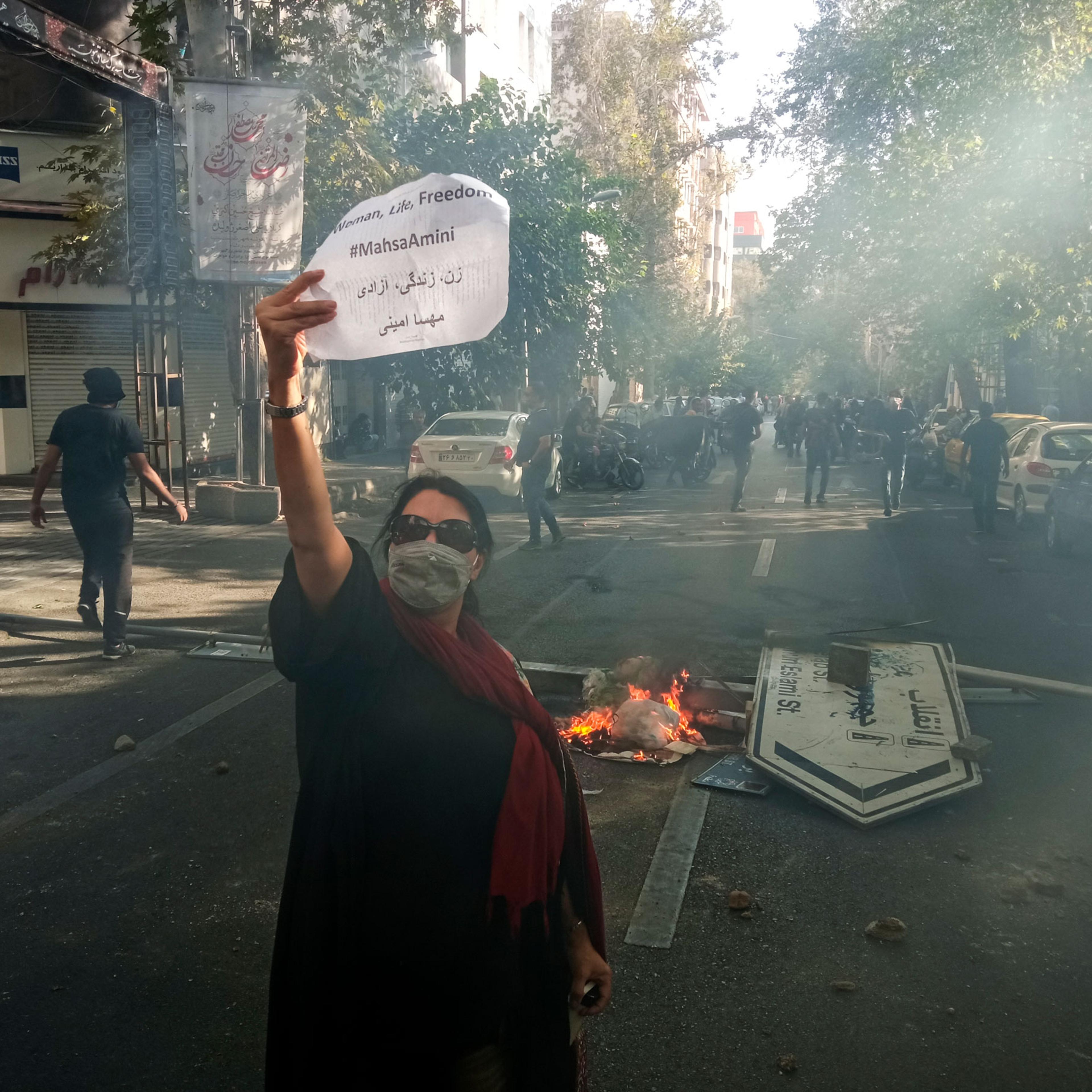 A woman holding the "Woman, Life, Freedom" sign, with the fallen street sign "Jomhouri Eslami" (Islamic Republic) in the background and tear gas in the air. October 1st 2022. Tehran, Iran.
