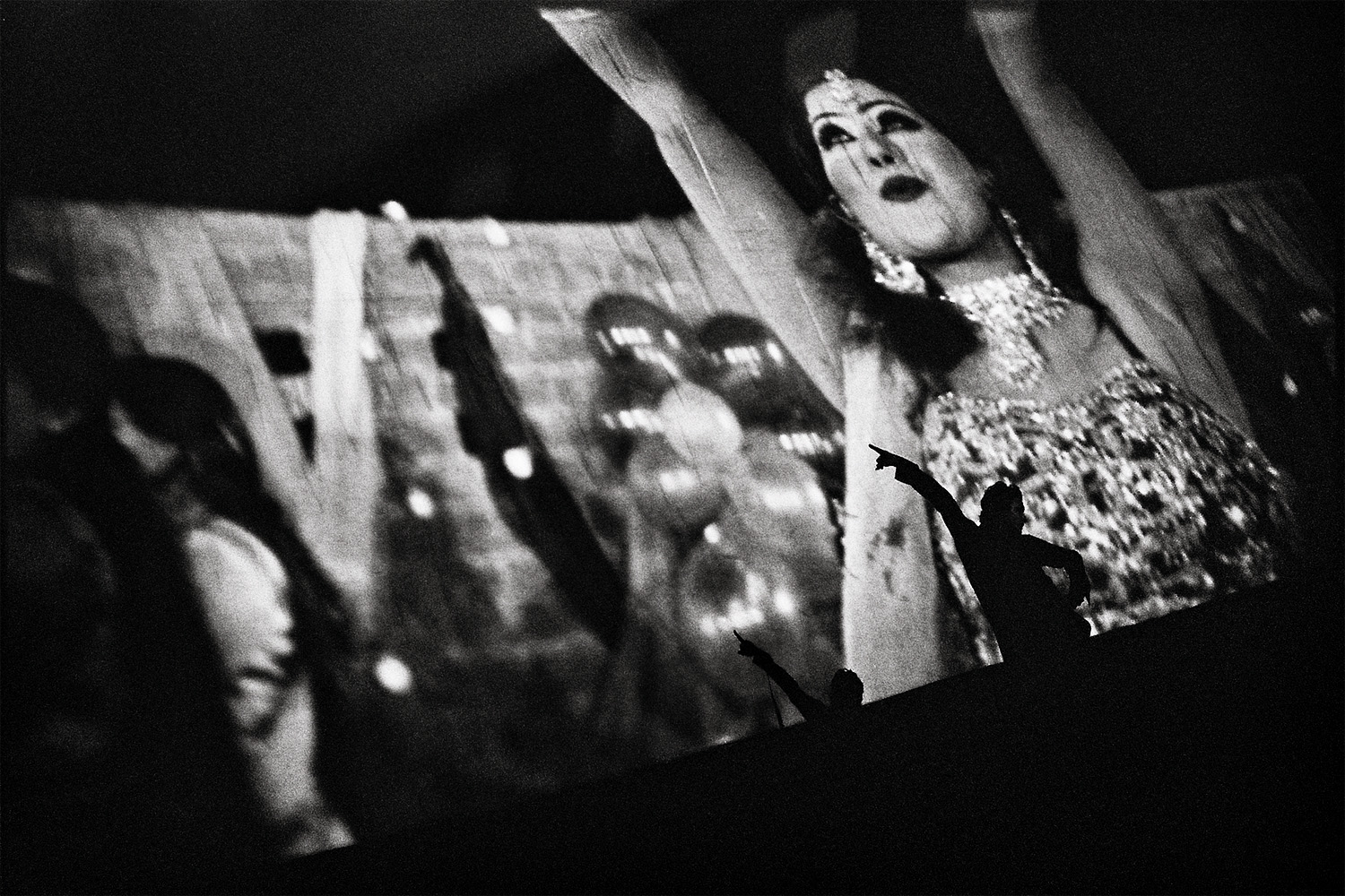 A Pashtun man dances on stage along to the musical number behind him on the screen. He is cheered on by the crowd. 