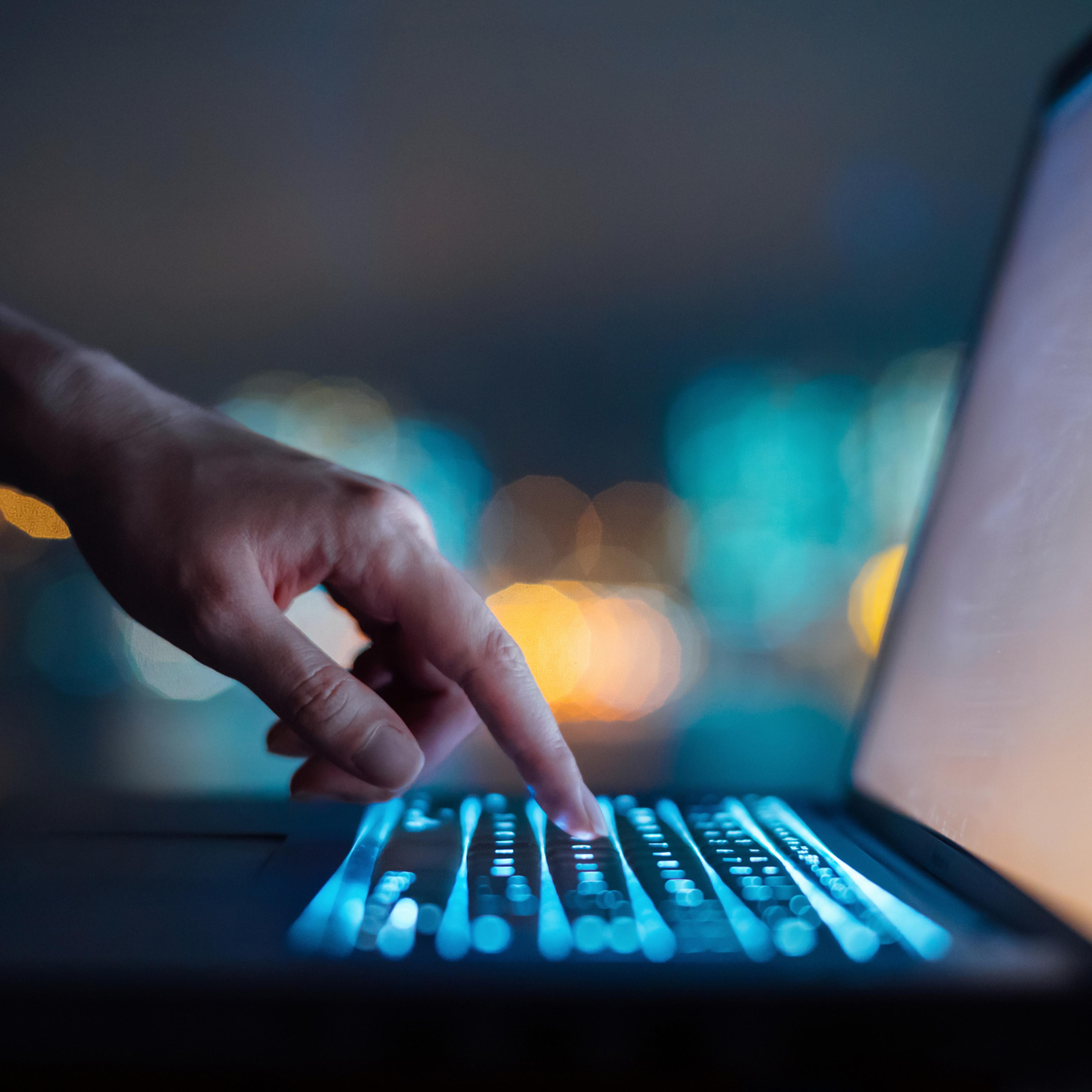 Close up of woman's hand typing on computer keyboard in the dark against colourful bokeh in background, working late on laptop at home
