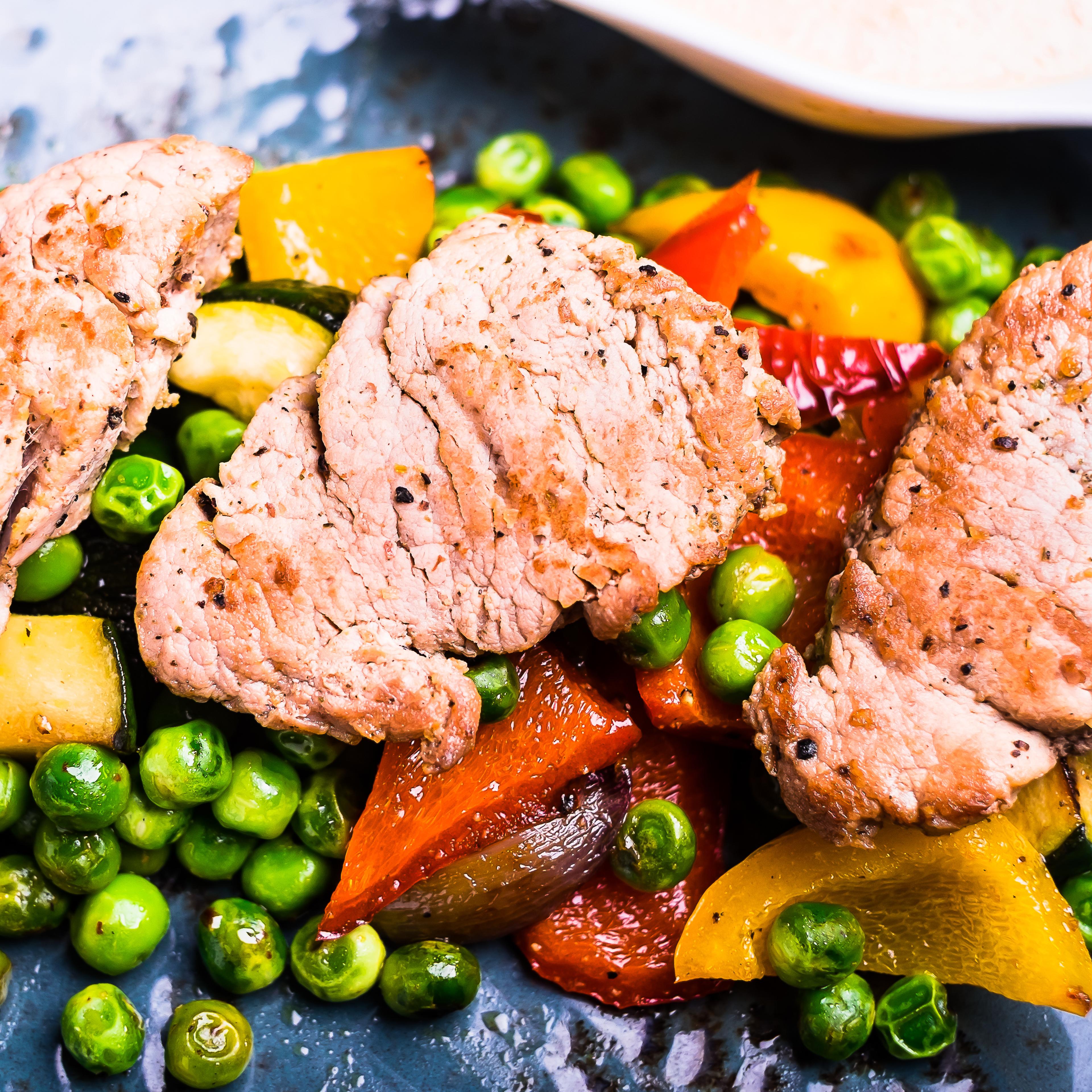 Roasted pork meat fillet chops with zucchini, onion, yellow and orange bell pepper, frozen peas on a plate with cream sauce, top view