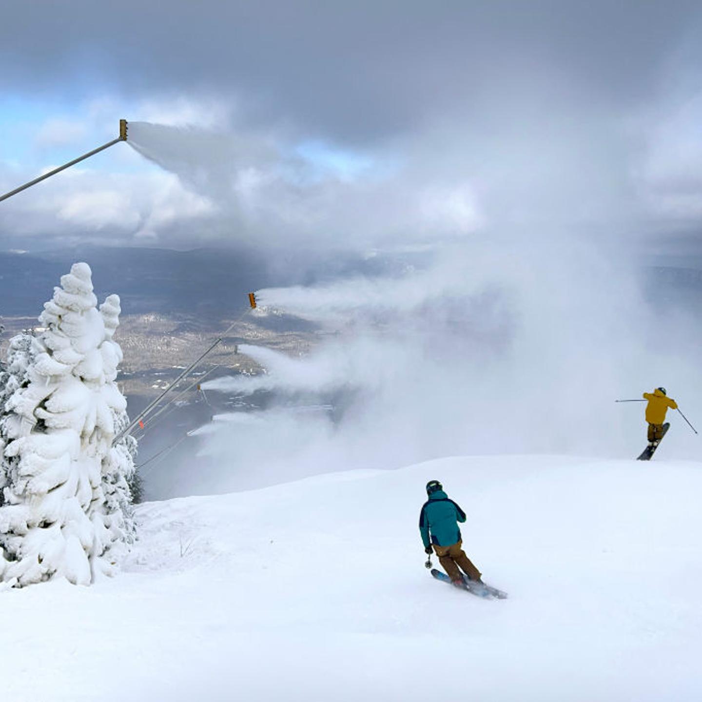Snow guns blow snow over skiers as they make their way down the hill.