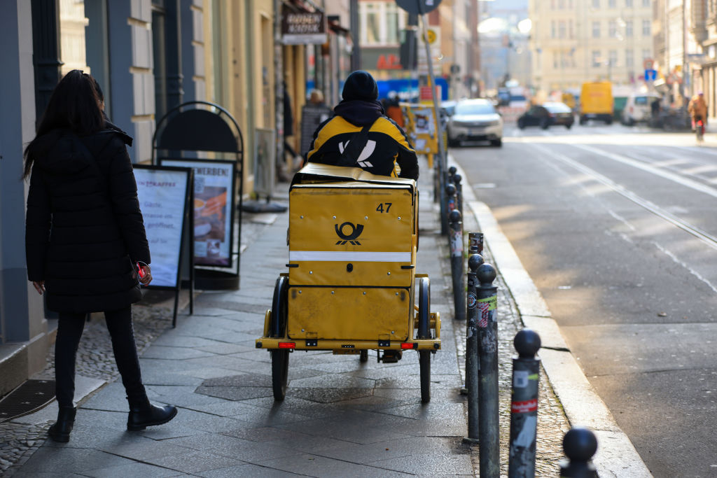 A delivery driver for Deutsche Post AG cycles on a sidewalk.
