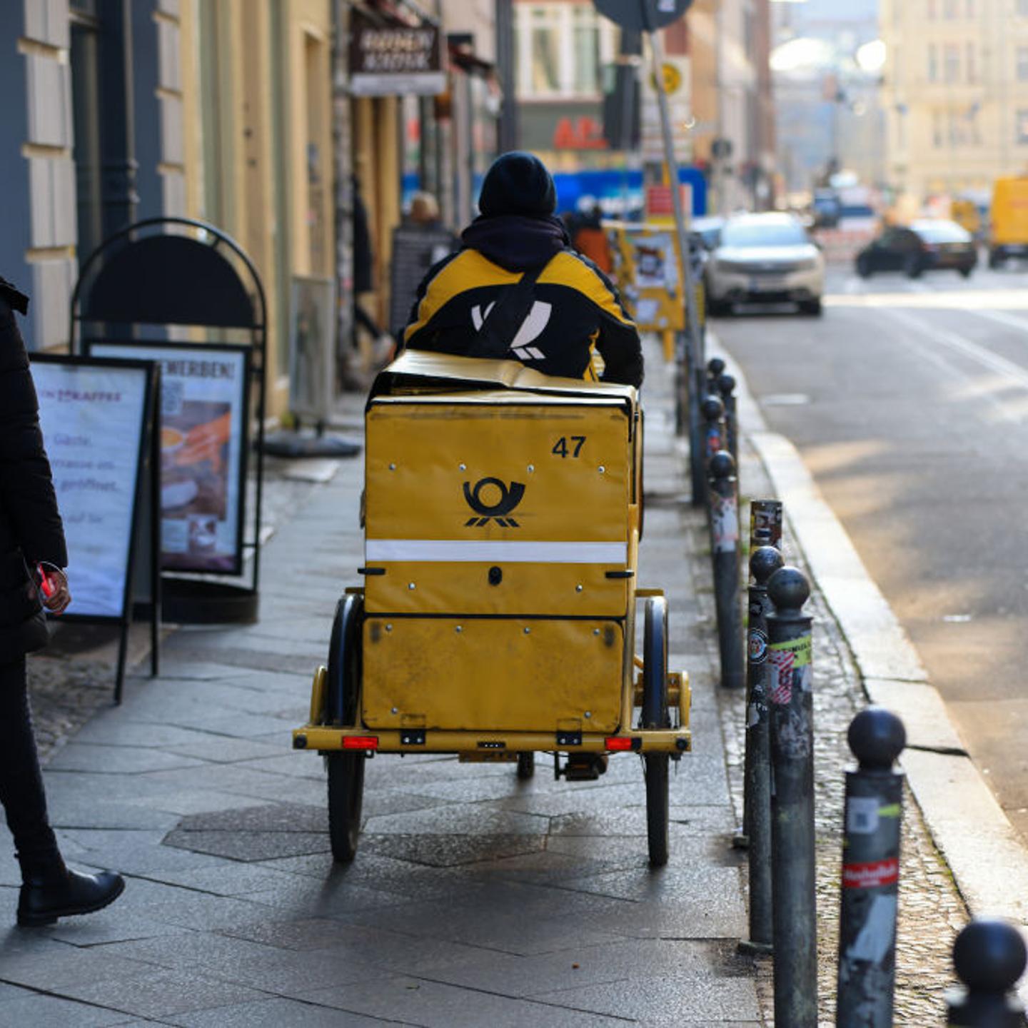 A delivery driver for Deutsche Post AG cycles on a sidewalk.