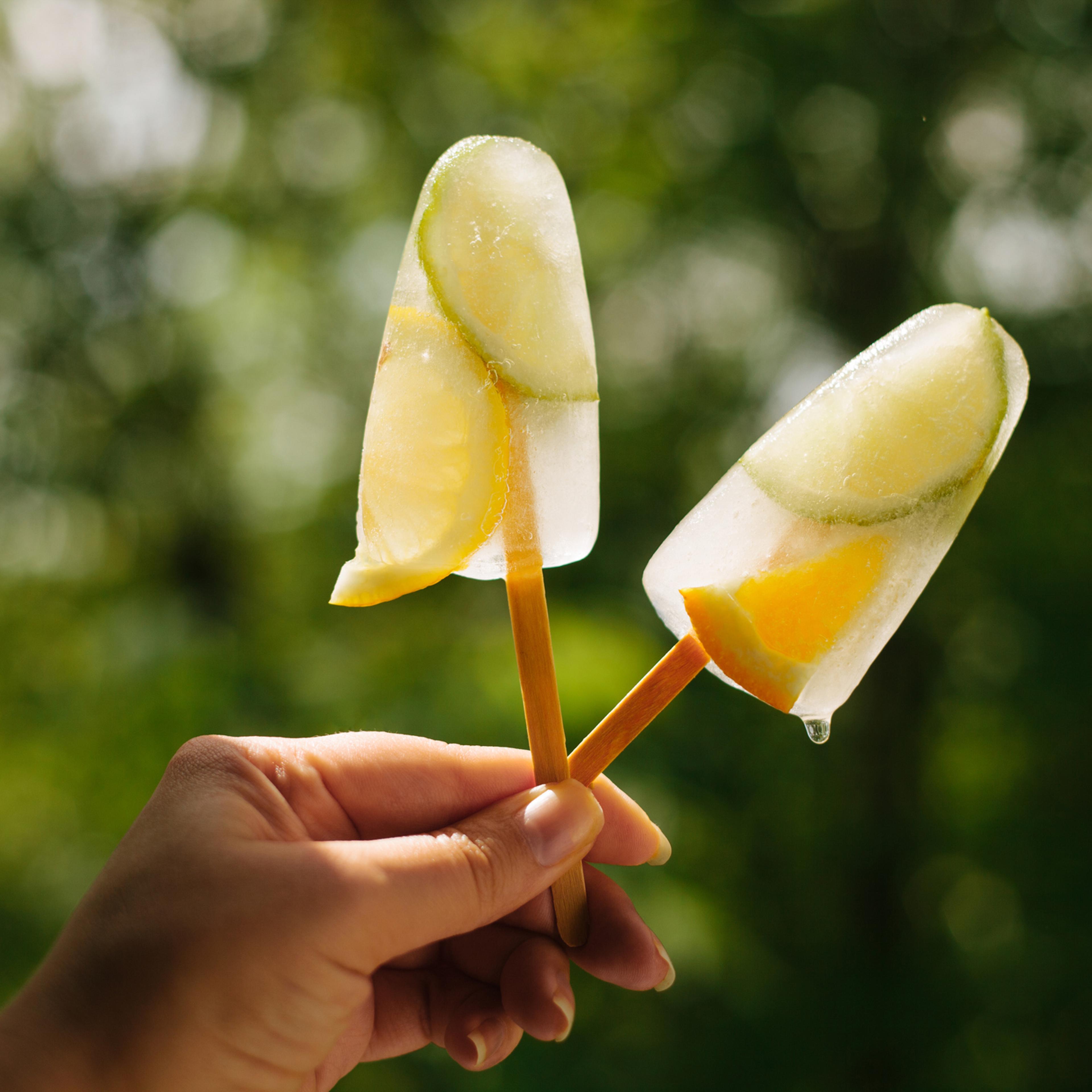 Two popsicles with lime and orange close-up in female hand against green background in bright sun light.