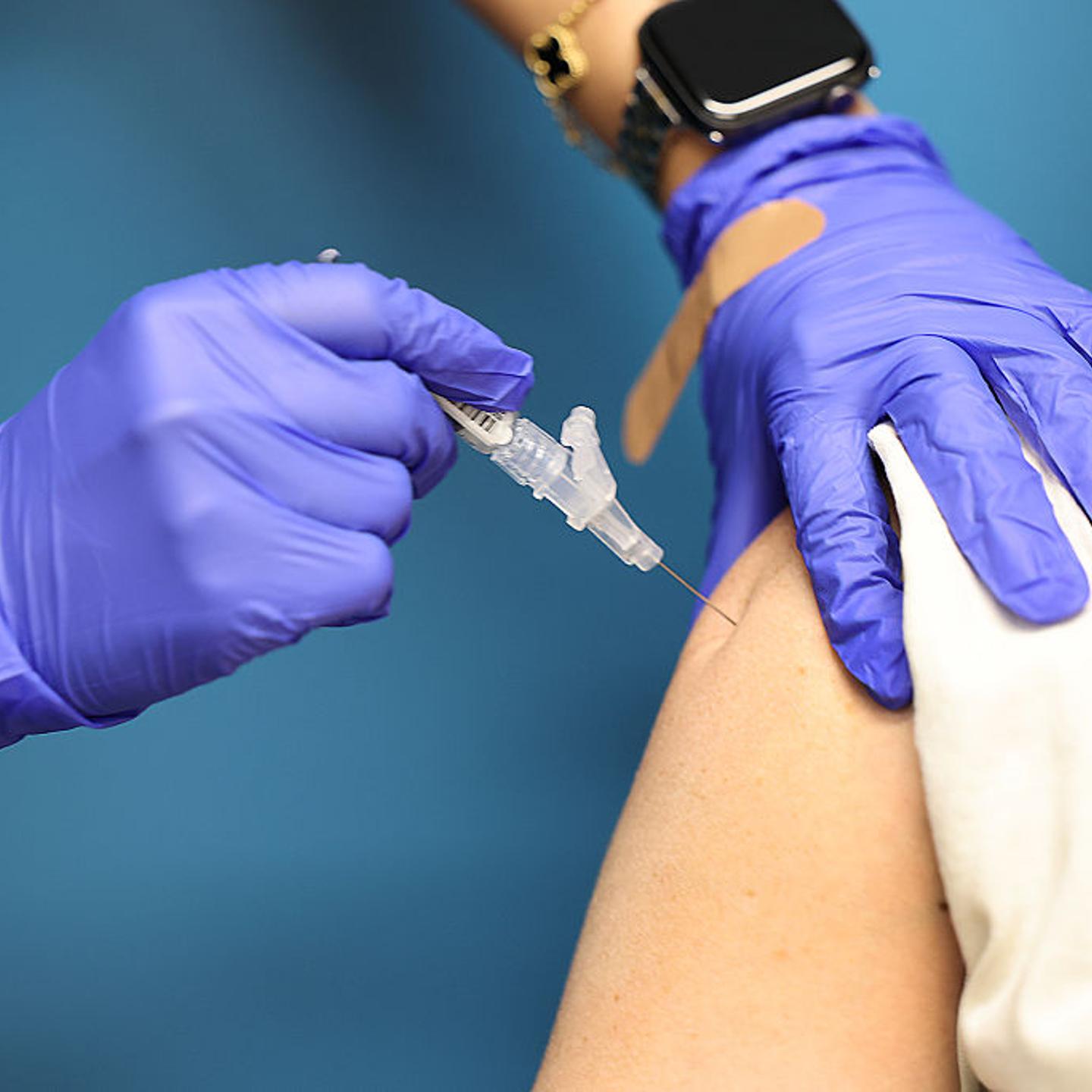 A medical professional with latex gloves inserts a vaccine into the shoulder of a patient with their sleeve rolled up.