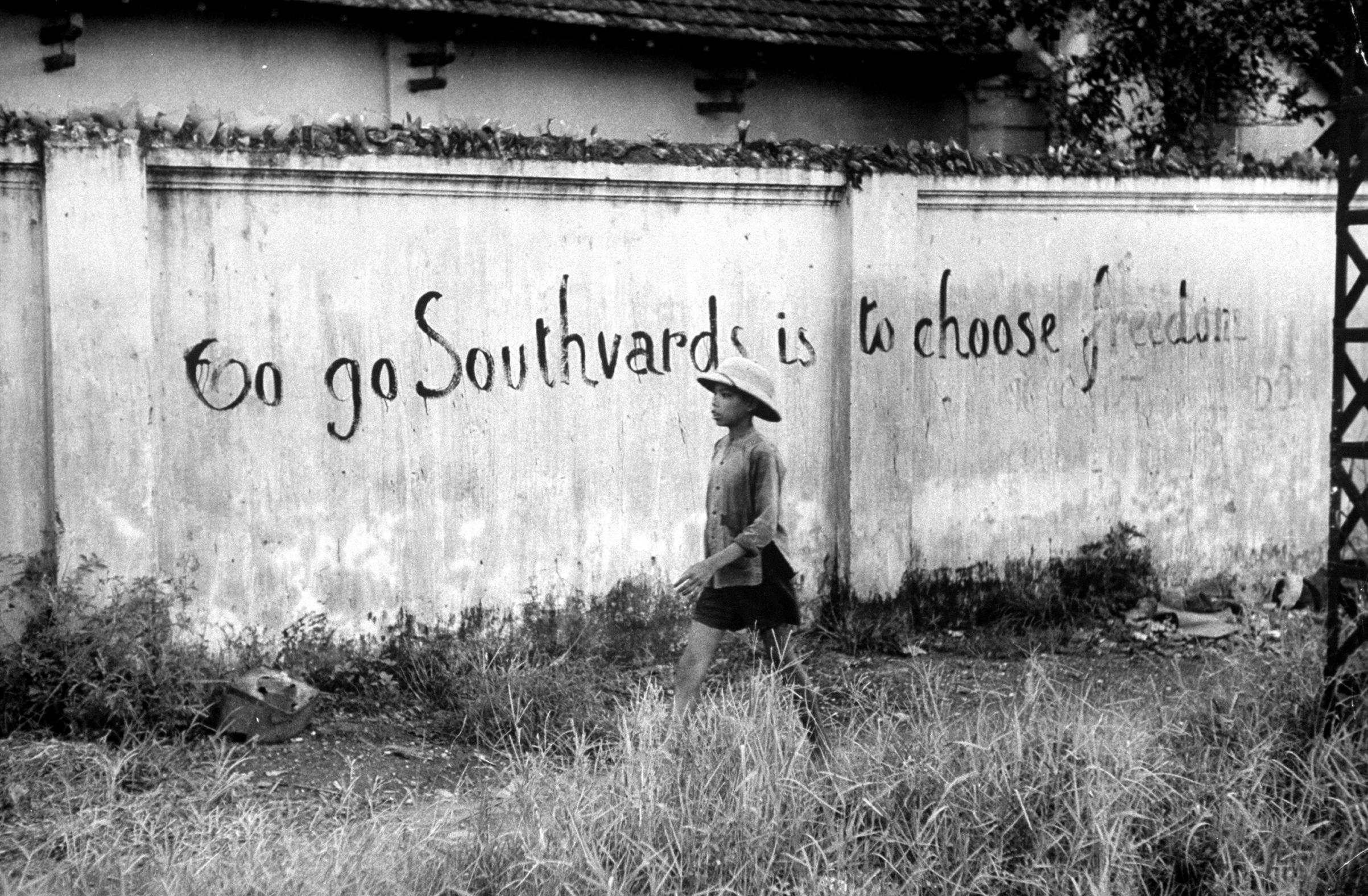 Boy walking past wall with writing that urges people to flee south from the Communists in Hanoi, Vietnam, 1954.