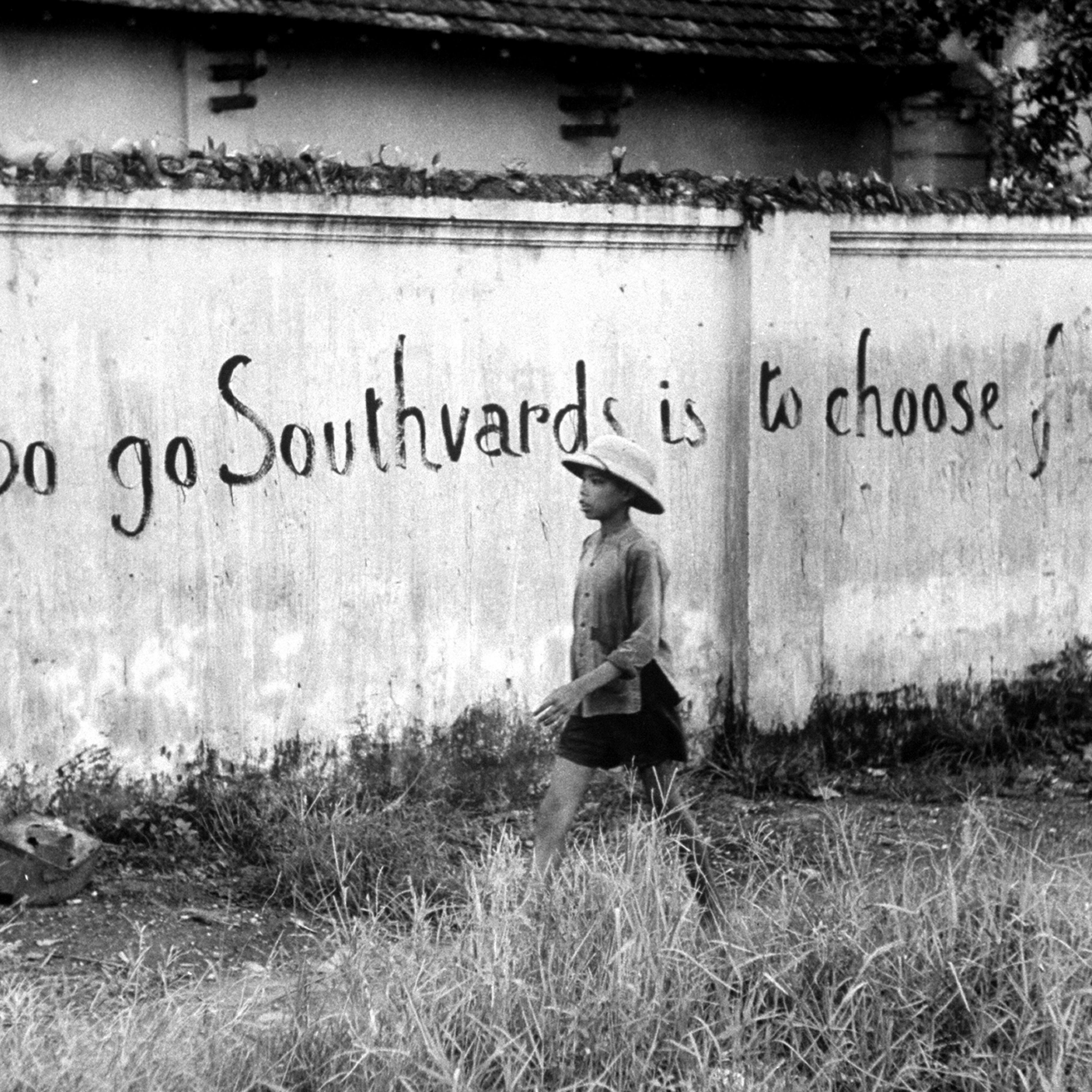 Boy walking past wall with writing that urges people to flee south from the Communists in Hanoi, Vietnam, 1954.